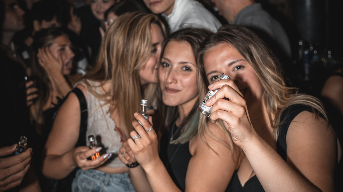 drei junge Frauen halten Fläschchen mit Cosmo Shot in der Hand, feiernde Personen im Hinergrund